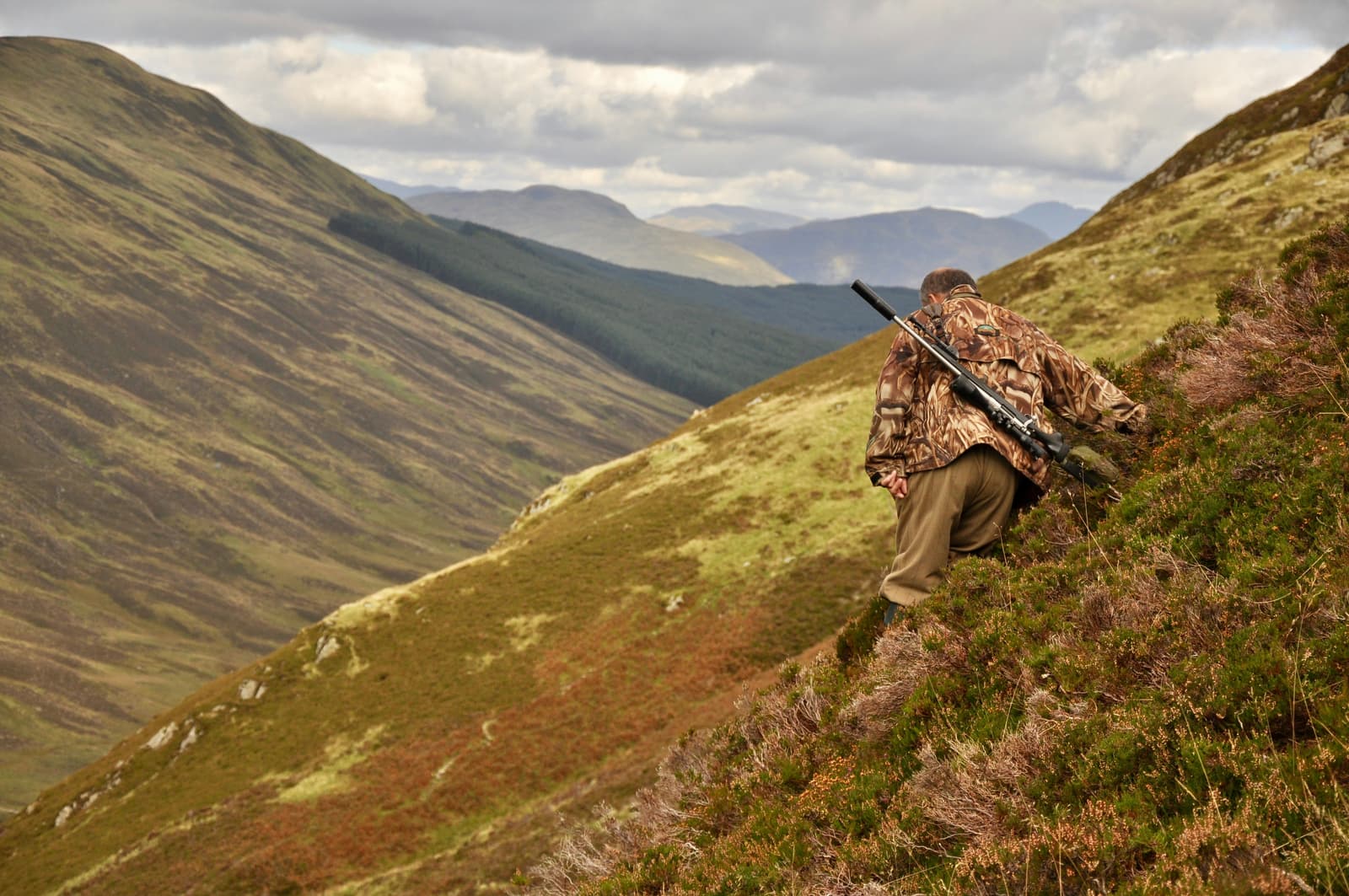 Hunter in camouflage sitting on a mountain hillside with rifle overlooking a valley
